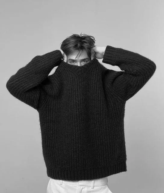 Black-and-white portrait of a person in an oversized chunky knit sweater pulled up toward the face and white trousers, suggesting comfort clothing as emotional shelter